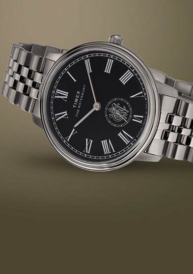 Timex Waterbury Metropolitan Sub-Second watch with polished stainless steel bracelet and black dial on a beige gradient background.