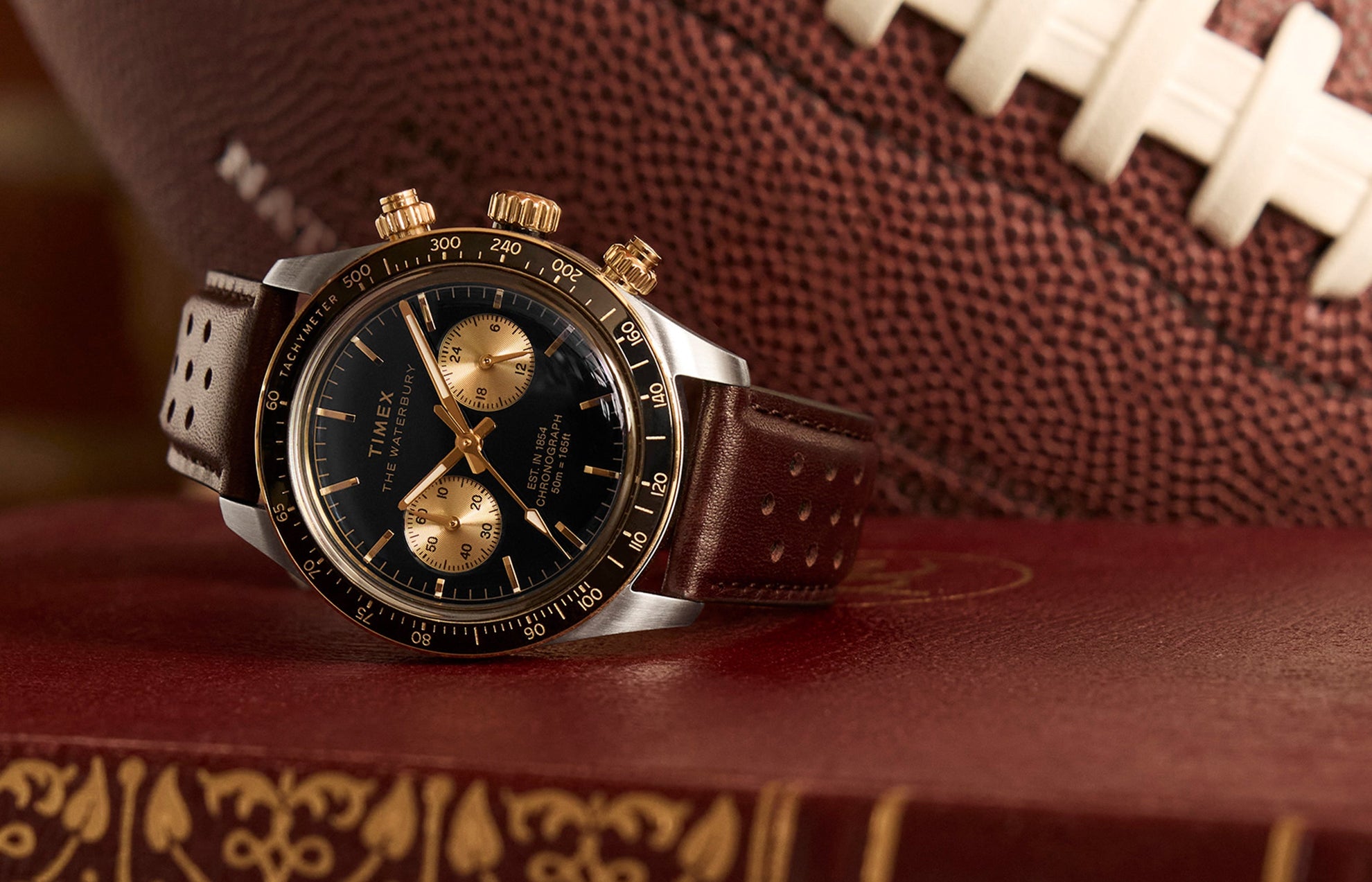 Timex Waterbury Heritage Chronograph with black dial, gold accents, and brown perforated leather strap resting on a vintage book with a football in the background.