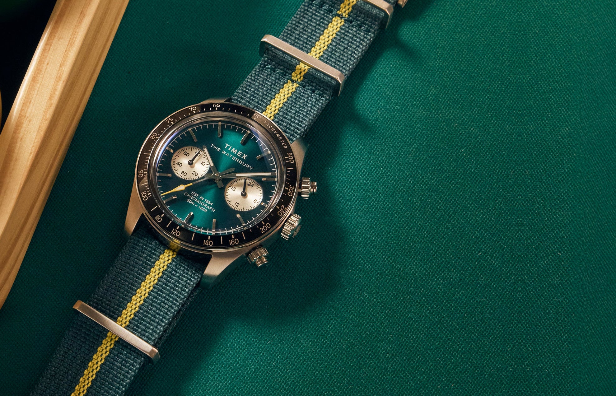 Timex Waterbury Heritage Chronograph with green sunray dial, silver sub-dials, and green fabric strap featuring a yellow center stripe resting on a billiard table.