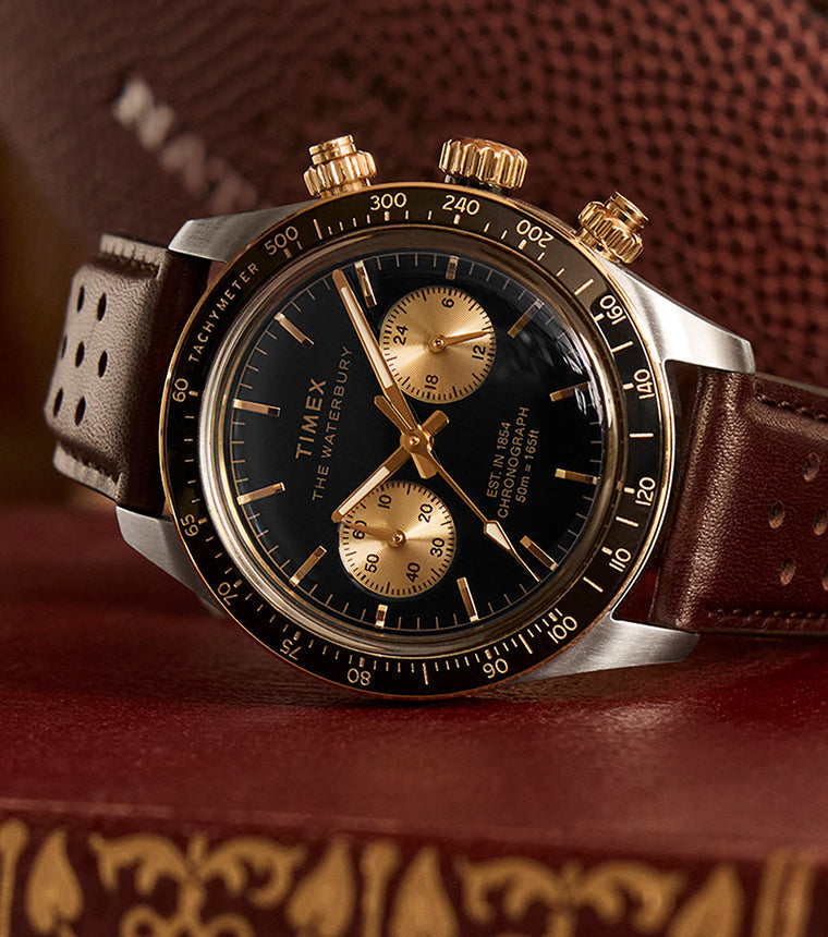 Timex Waterbury Heritage Chronograph with black dial, gold accents, and brown perforated leather strap resting on a vintage book with a football in the background.