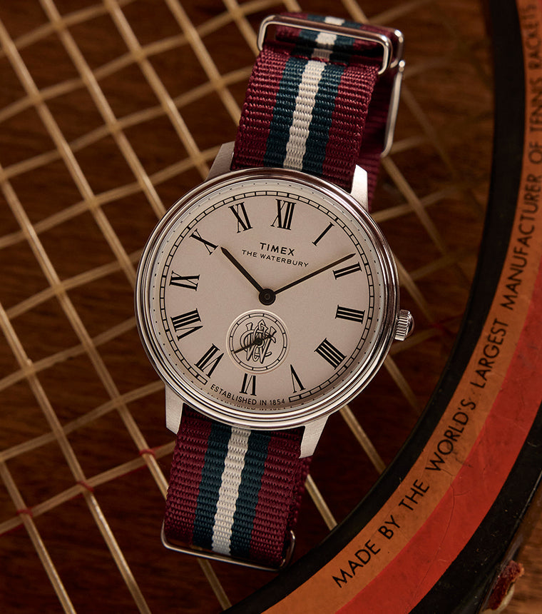 Timex Waterbury Metropolitan Sub-Second with white dial and red, white, and blue-striped fabric strap placed on a vintage wooden tennis racket.