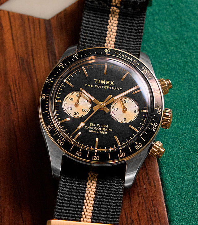 Timex Waterbury Heritage Chronograph with black dial, gold-tone accents, and black fabric strap featuring a tan center stripe resting on a wooden pool table.