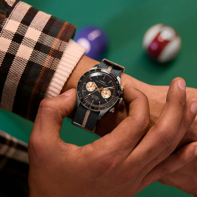 Close-up of the Timex Waterbury Heritage Chronograph on wrist, showing black and gold dial with striped fabric strap beside pool balls.