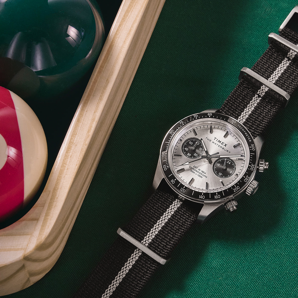 Timex Waterbury Heritage Chronograph positioned beside billiard balls, showcasing its silver dial, black sub-dials, and sporty design.