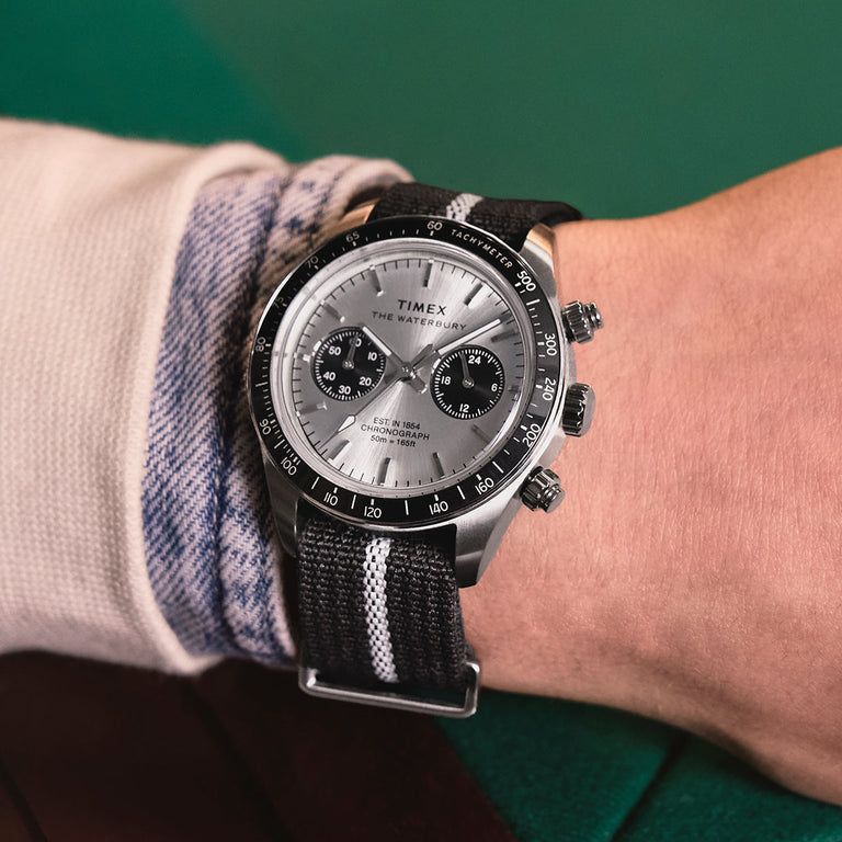 Timex Waterbury Heritage Chronograph with silver sunray dial, black sub-dials, and black fabric strap featuring a white center stripe worn on wrist.