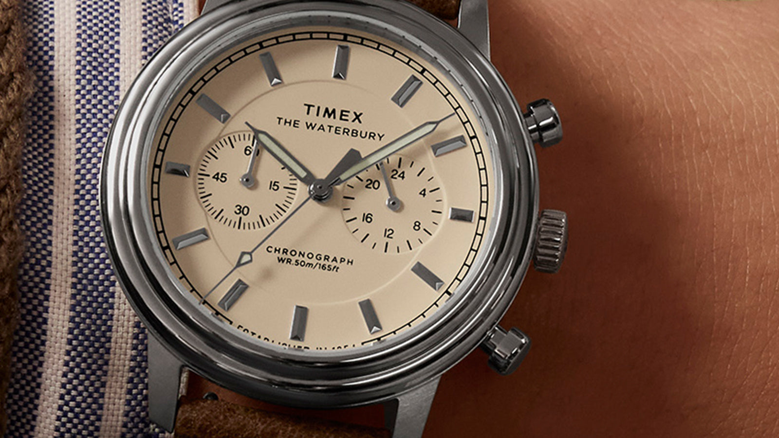 Timex Waterbury Metropolitan Chronograph worn on wrist, showing cream dial, chronograph sub-dials, and brown leather strap paired with denim and knit layers.