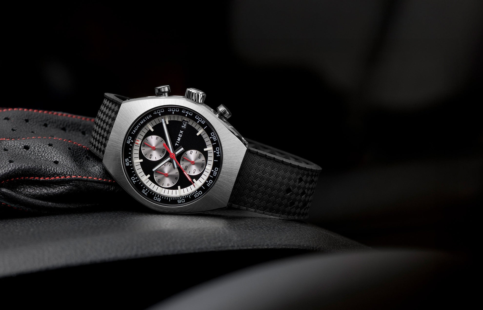 TIMEX® Legacy Tonneau Chronograph watch with black dial, silver-tone case, and textured black strap resting on perforated leather with red stitching.