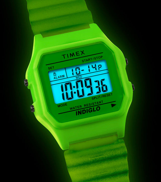 TIMEX® T80 digital watch illuminated with bright green INDIGLO® backlight in a dark setting, showing the display clearly glowing blue.