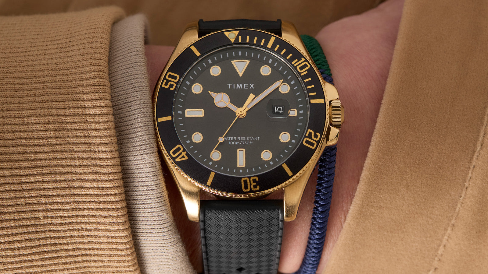 Timex Harborside Coast watch with a black dial and gold-tone case worn on the wrist with a tan coat sleeve and blue-green woven bracelet.