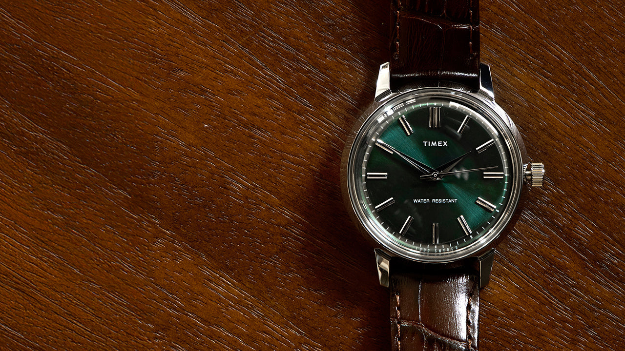 Todd Snyder x Timex Olive Marlin Automatic watch with a green sunray dial and brown leather strap, displayed flat on a textured wooden surface.