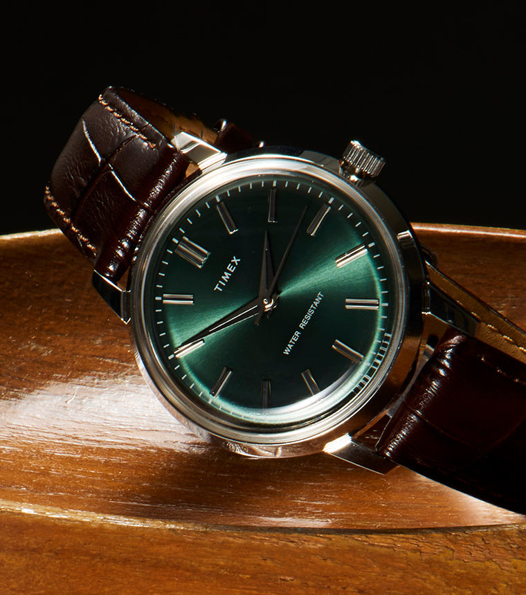 Todd Snyder x Timex Olive Marlin Automatic watch with a green dial resting at an angle on a polished wooden pedestal against a dark background.