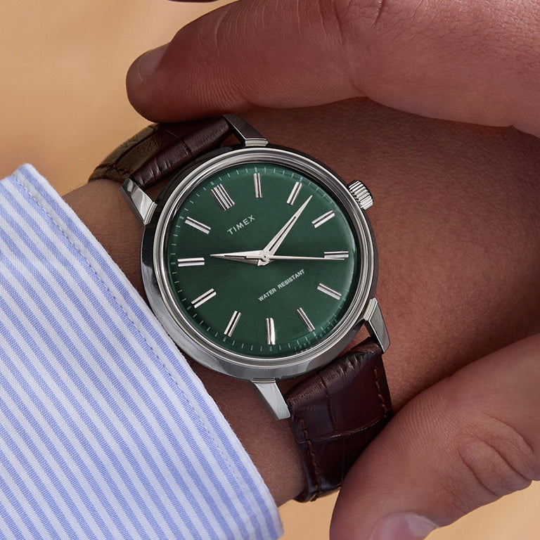 Todd Snyder x Timex Olive Marlin Automatic worn on the wrist, featuring a green dial and brown leather strap paired with a blue striped dress shirt.