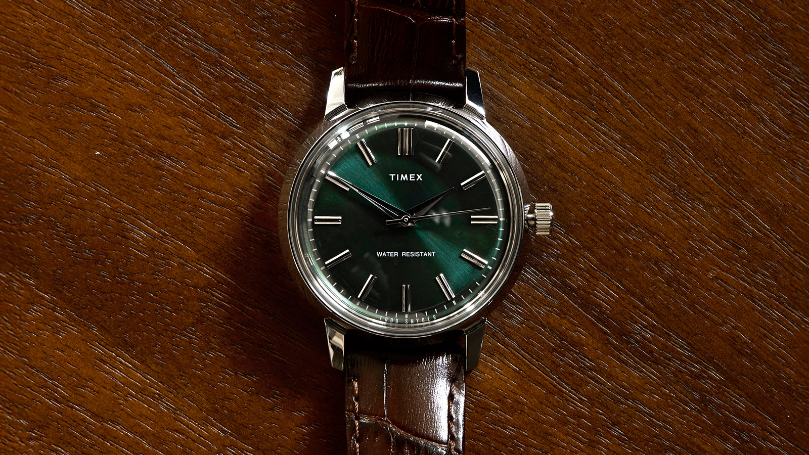 Front-facing view of the Todd Snyder x Timex Olive Marlin Automatic, showing its green dial, silver markers, and brown leather strap on a wood background.