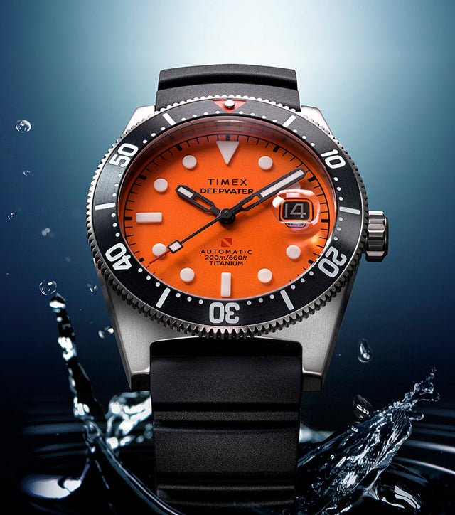 Timex Deepwater Reef 200 watch with a bright orange dial emerging from dark water, with droplets splashing around the titanium case and black dive bezel.