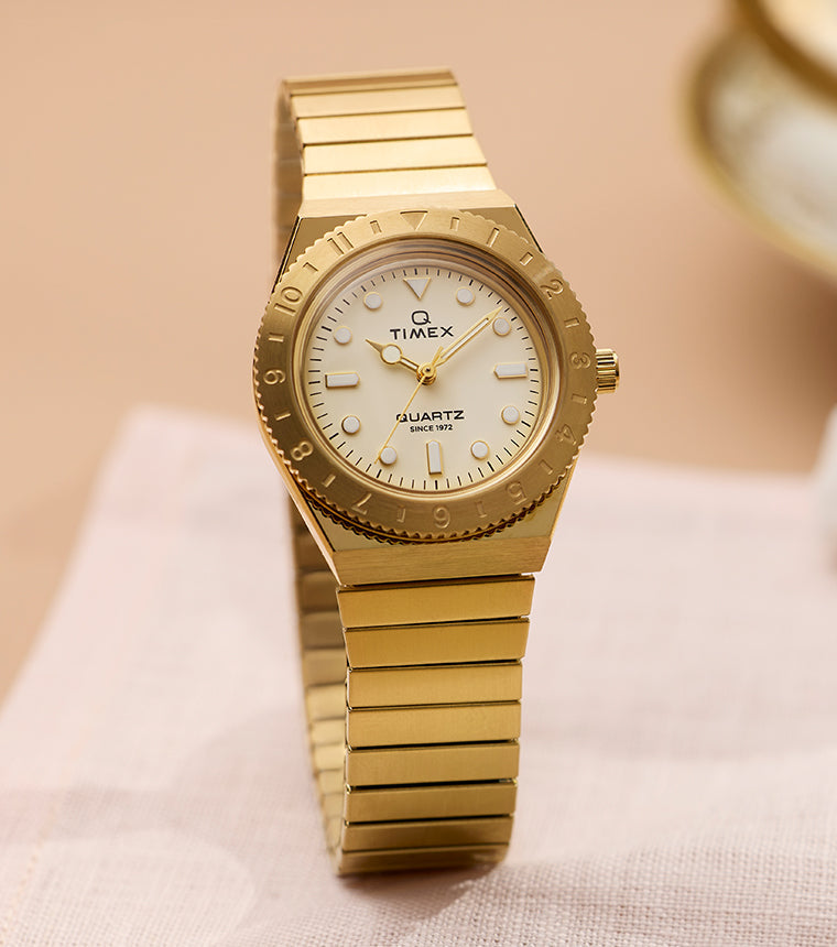 Q Timex® Mini gold-tone watch featuring a cream dial, rotating bezel, and gold expansion bracelet, displayed on a soft neutral tabletop.