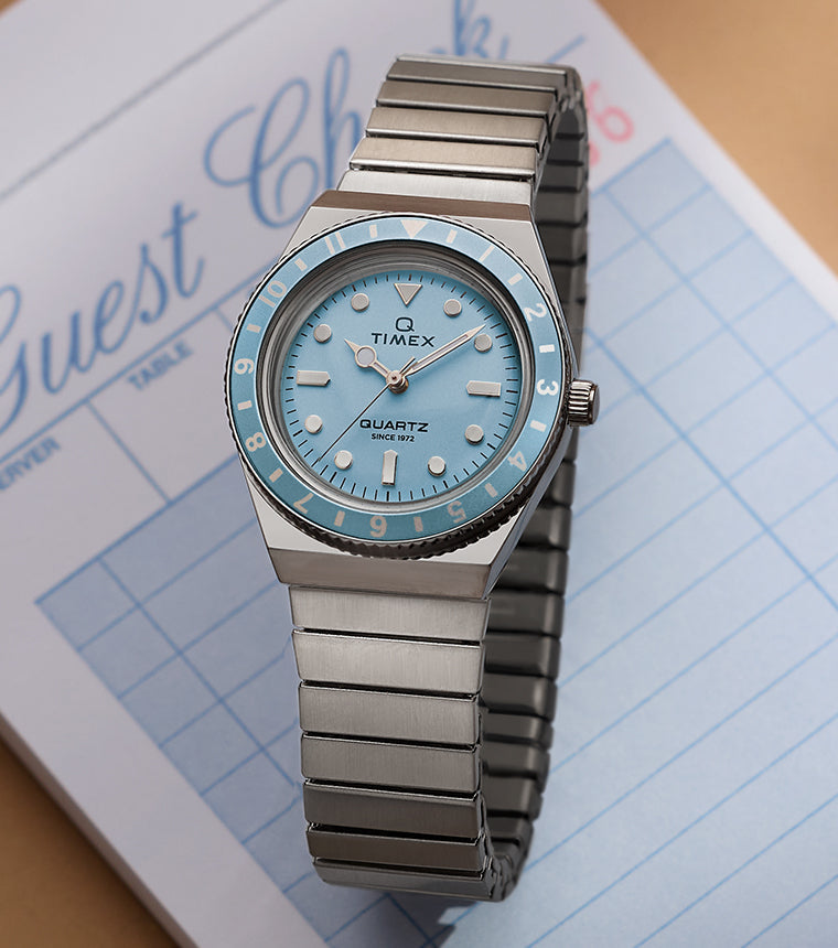 Q Timex® Mini silver-tone watch with light blue dial, rotating bezel, and stainless steel expansion bracelet, styled on a guest check pad.