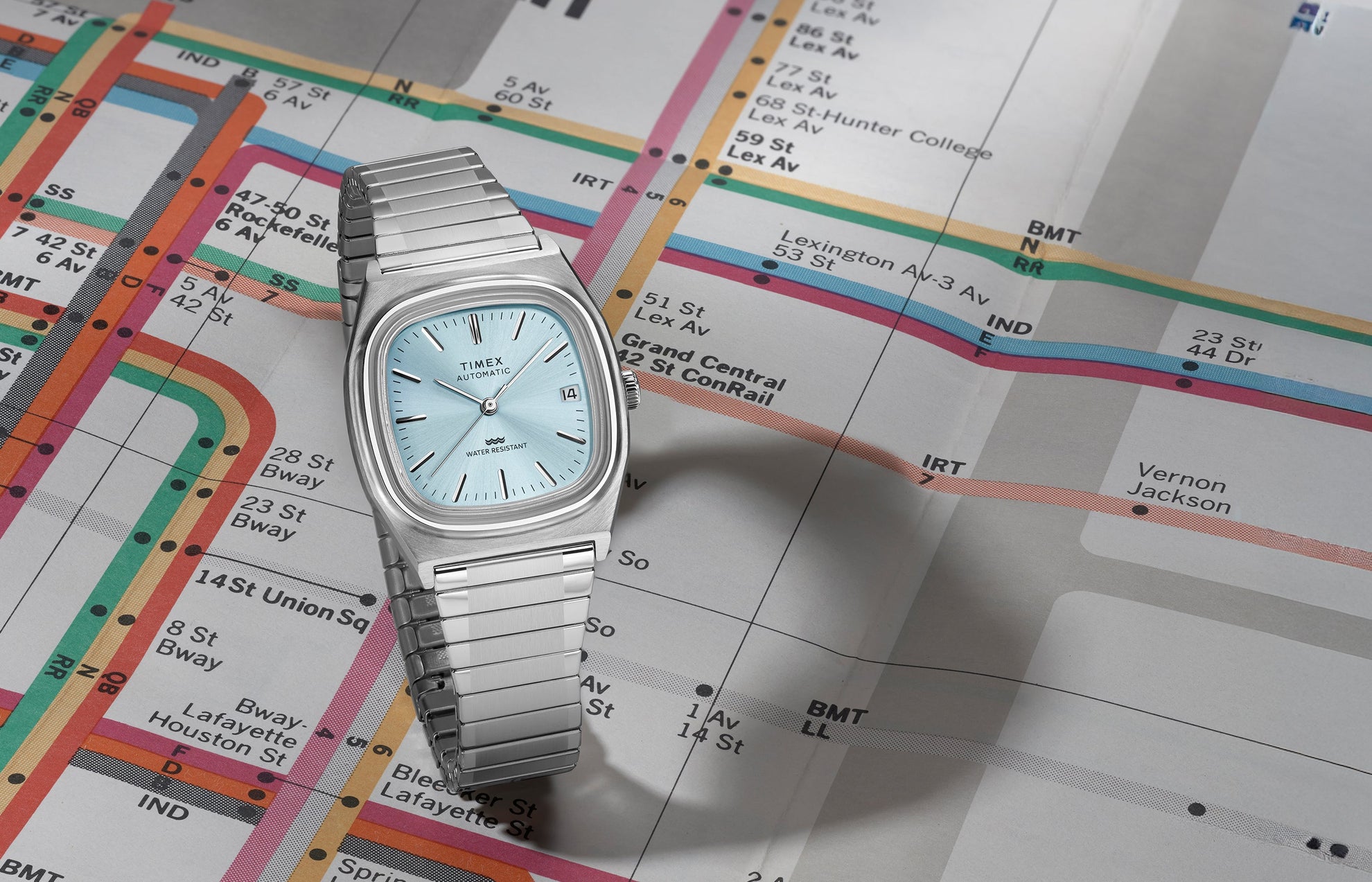Timex® Automatic 1983 E Line stainless steel watch with light blue sunray dial displayed at an angle on a vintage subway map.