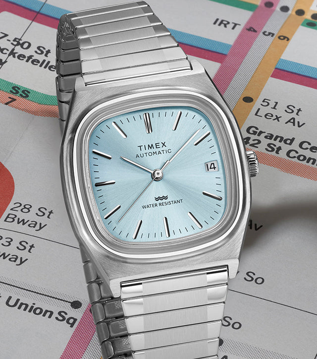 Timex® Automatic 1983 E Line stainless steel watch with light blue sunray dial displayed at an angle on a vintage subway map.