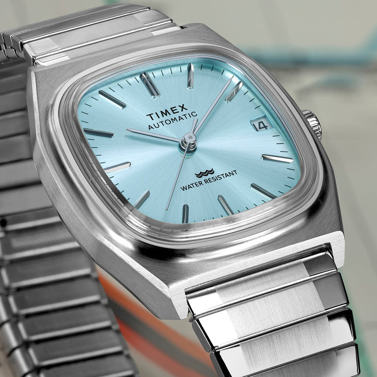 Close-up of the Timex® Automatic 1983 E Line watch showing a light blue sunray dial, silver hour markers, date window, and brushed stainless steel case.
