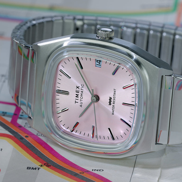 Angled view of the Timex® Automatic 1983 E Line watch highlighting the pink dial, polished crown, and stainless steel expansion bracelet on a subway map.