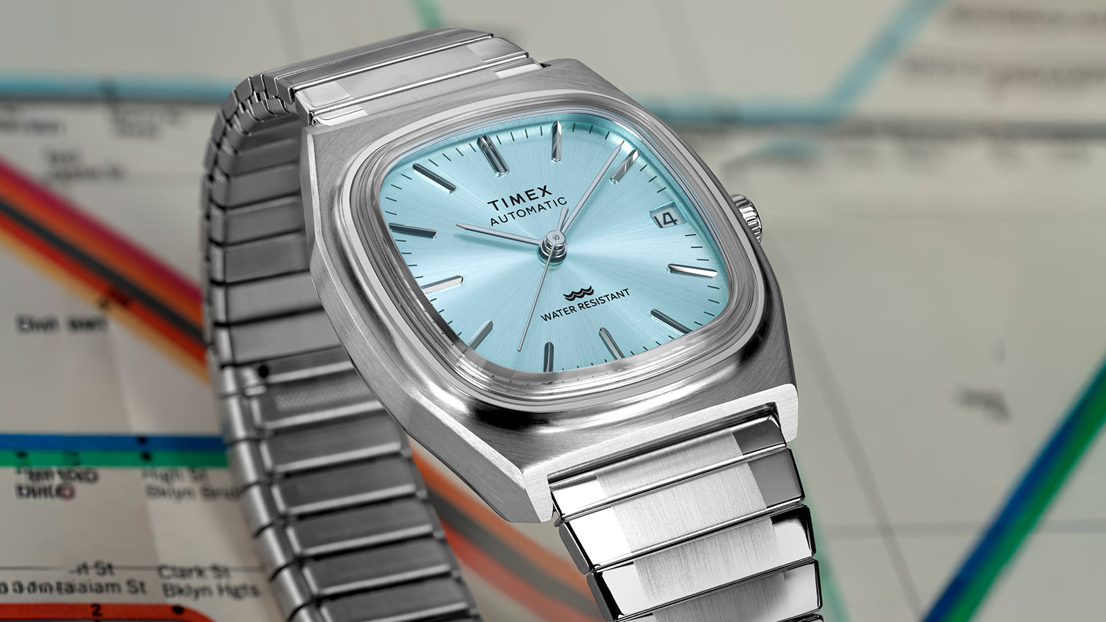 Angled close-up of the Timex® Automatic 1983 E Line watch emphasizing the brushed stainless steel case, light blue dial, and metal expansion bracelet.