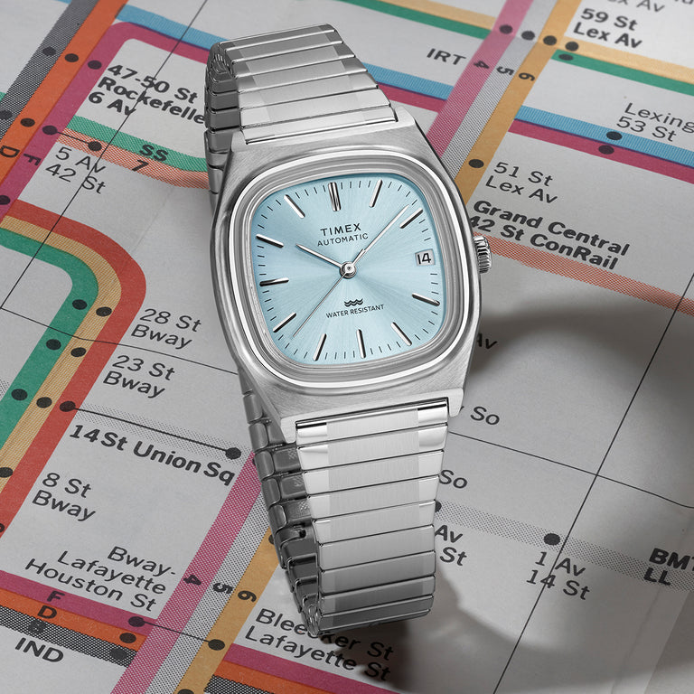Timex E Line Automatic watch with light blue dial and stainless steel bracelet displayed on a colorful NYC subway map.