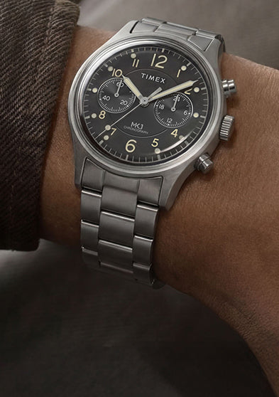 Timex MK1 Chronograph stainless steel watch on wrist with black dial, luminous Arabic numerals, and brushed metal bracelet.