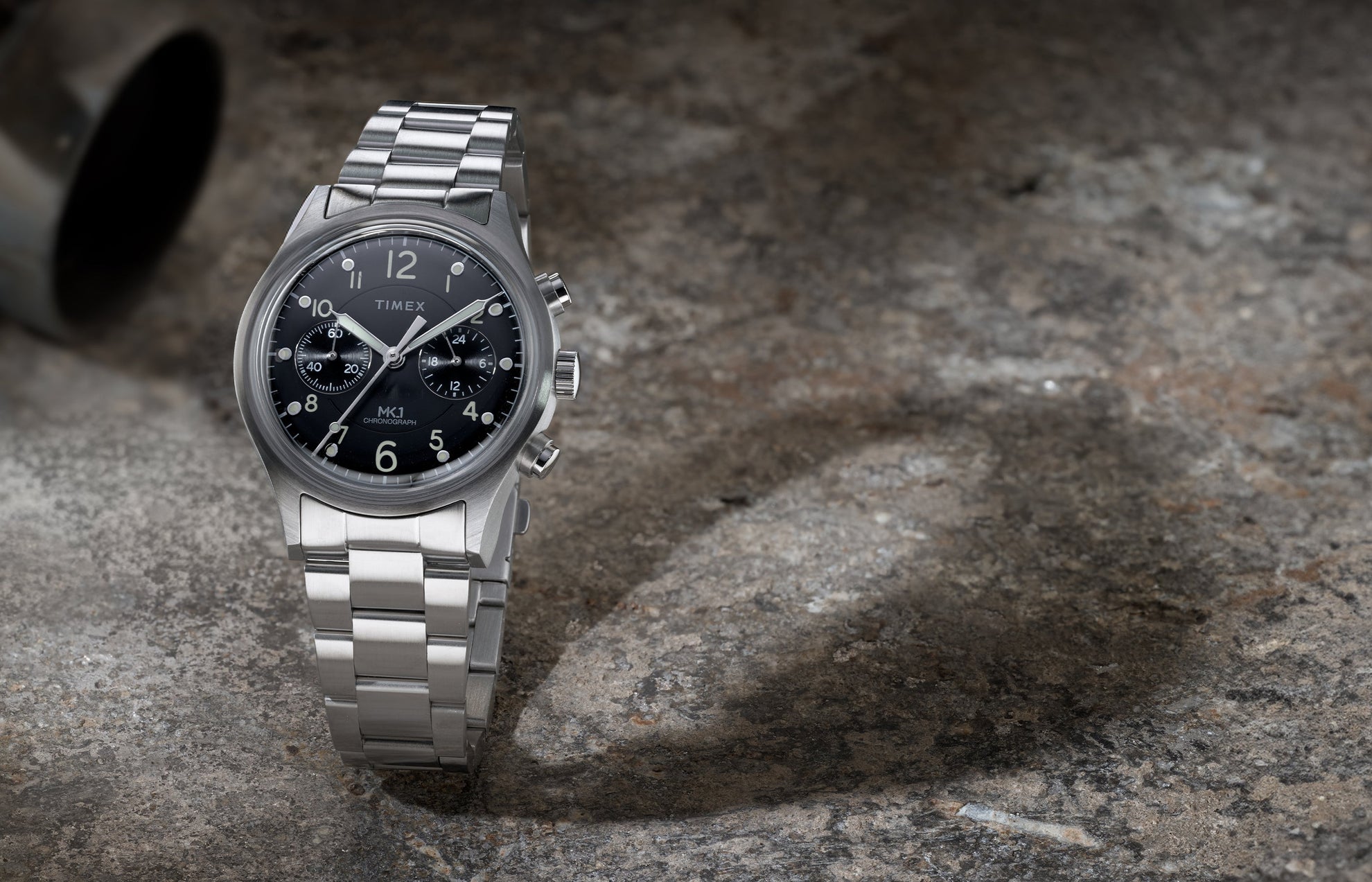 Timex MK1 Chronograph with stainless steel bracelet and black dial, shown standing upright on a textured stone surface.