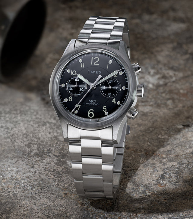 Timex MK1 Chronograph with stainless steel bracelet and black dial, shown standing upright on a textured stone surface.