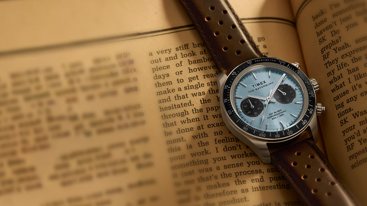 Timex Waterbury Heritage Chronograph with brown leather strap displayed on an open book, showcasing the light blue dial and tachymeter bezel.