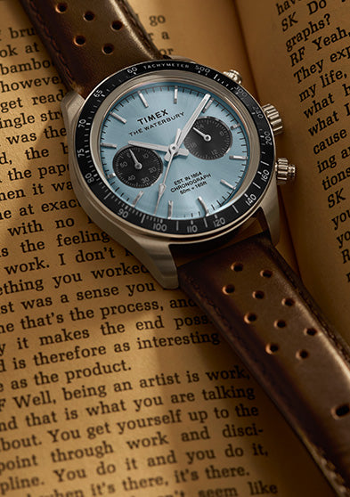 Timex Waterbury Heritage Chronograph with brown leather strap displayed on an open book, showcasing the light blue dial and tachymeter bezel.