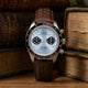 Timex Waterbury Heritage Chronograph with brown leather strap and light blue dial, shown front-facing against a vintage backdrop.