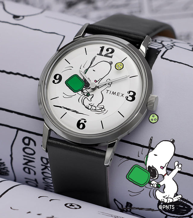 Peanuts® x Timex Marlin® Pickleball watch with black leather strap and white dial featuring Snoopy playing pickleball, displayed on Peanuts comic strip background.