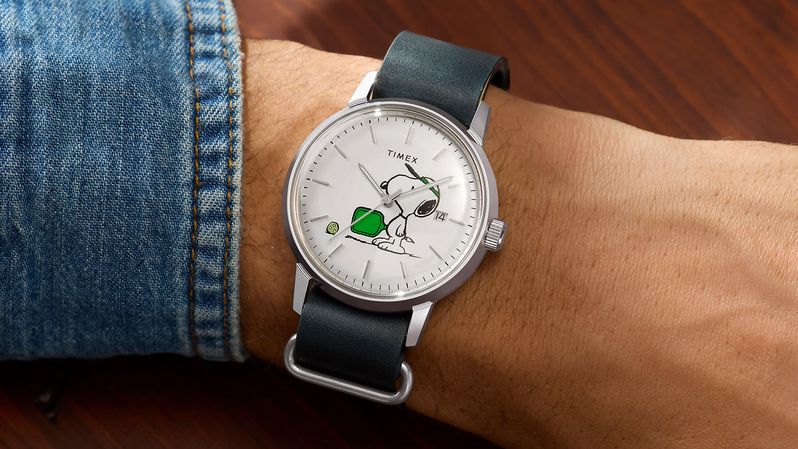 Peanuts® x Timex Marlin® Automatic Pickleball watch worn on wrist, featuring Snoopy playing pickleball on a white dial, stainless-steel case, date window, and dark green leather strap.