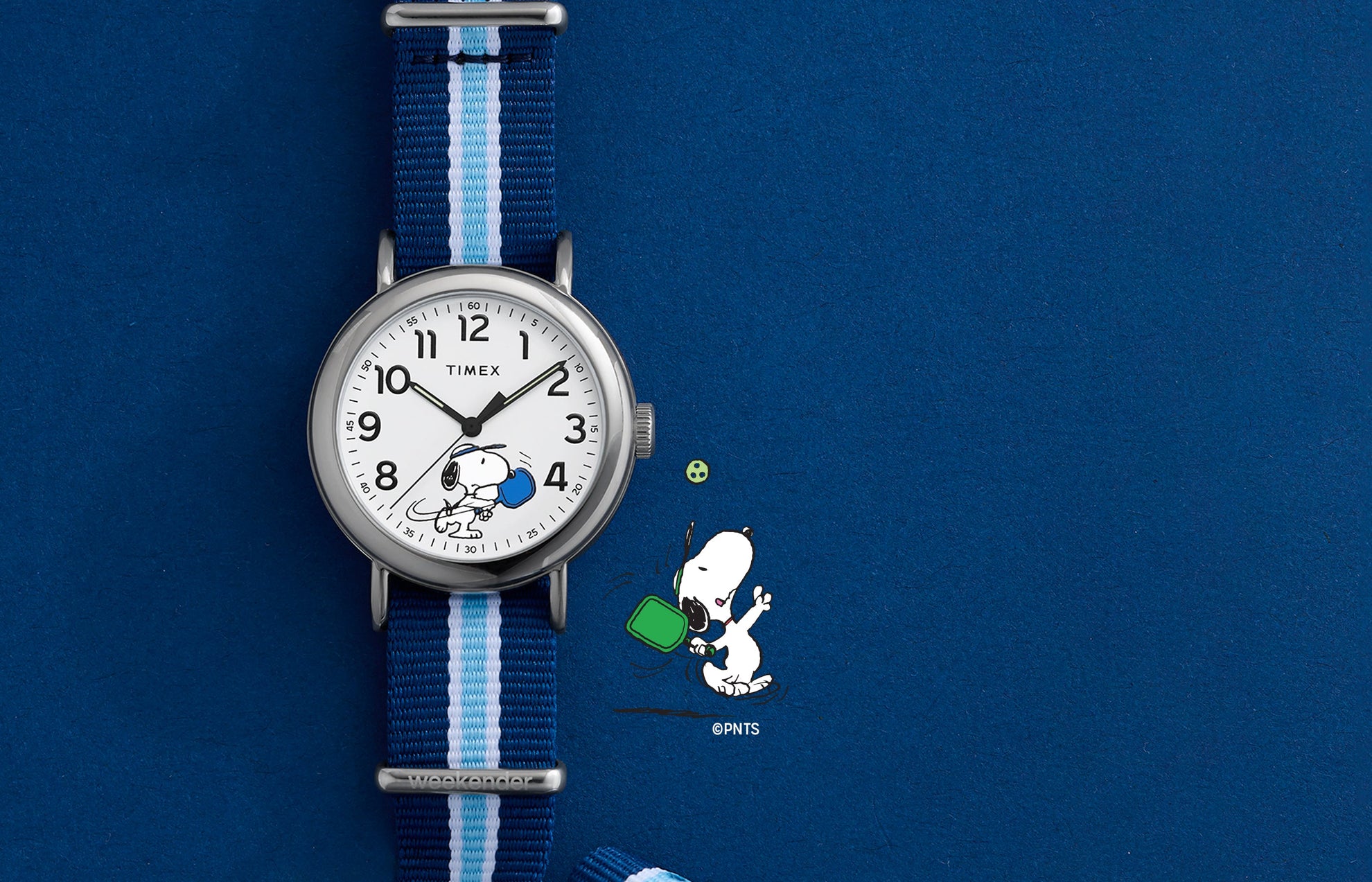 Peanuts® x Timex Weekender® Pickleball watch with white dial, full Arabic numerals, and blue striped fabric slip-thru strap displayed on blue background.