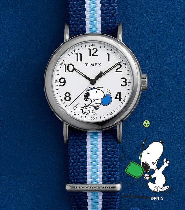 Peanuts® x Timex Weekender® Pickleball watch with white dial, full Arabic numerals, and blue striped fabric slip-thru strap displayed on blue background.