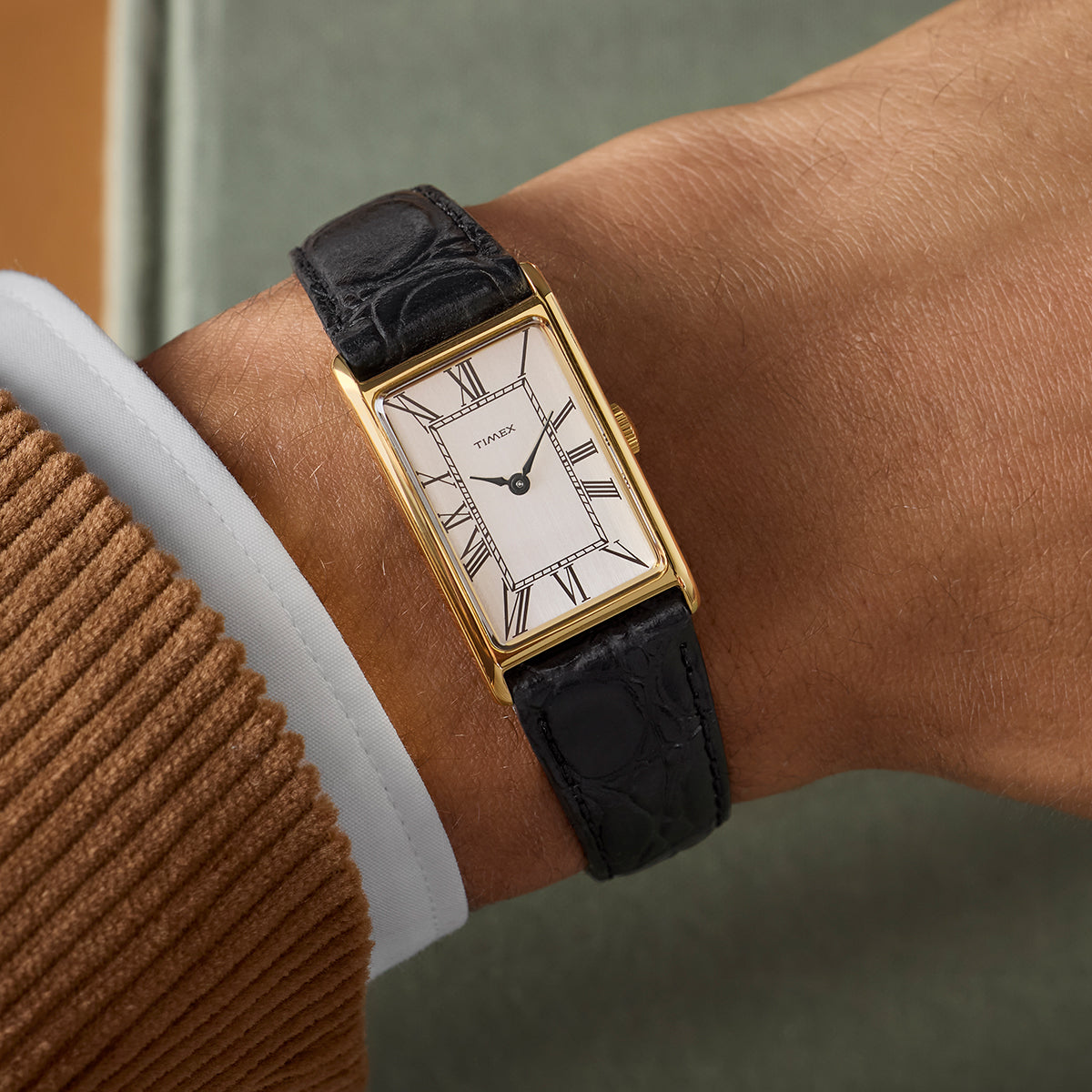 Gold-tone rectangular Timex 1976 Lexington Reissue watch with a silver dial and black Roman numerals, worn on a wrist with a black embossed leather strap.