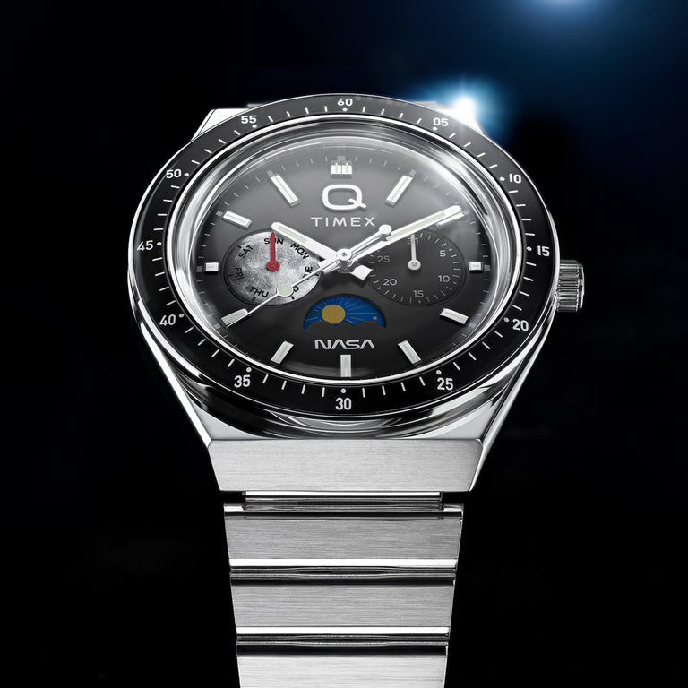 Close-up of Q Timex® x NASA watch featuring brushed stainless steel bracelet, black bezel, gray dial with day subdial, chronograph-style subdial, and NASA logo at 6 o’clock.