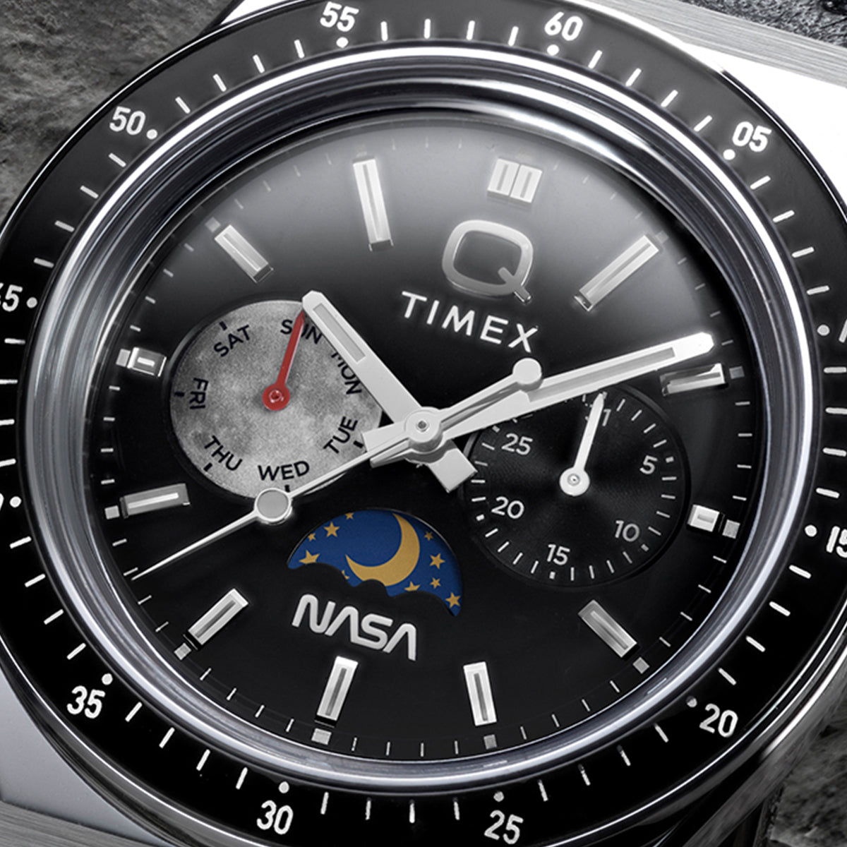 Close-up of Q Timex® x NASA black dial featuring day-of-week subdial with red hand, secondary subdial, moonphase detail, luminous markers, and NASA logo.
