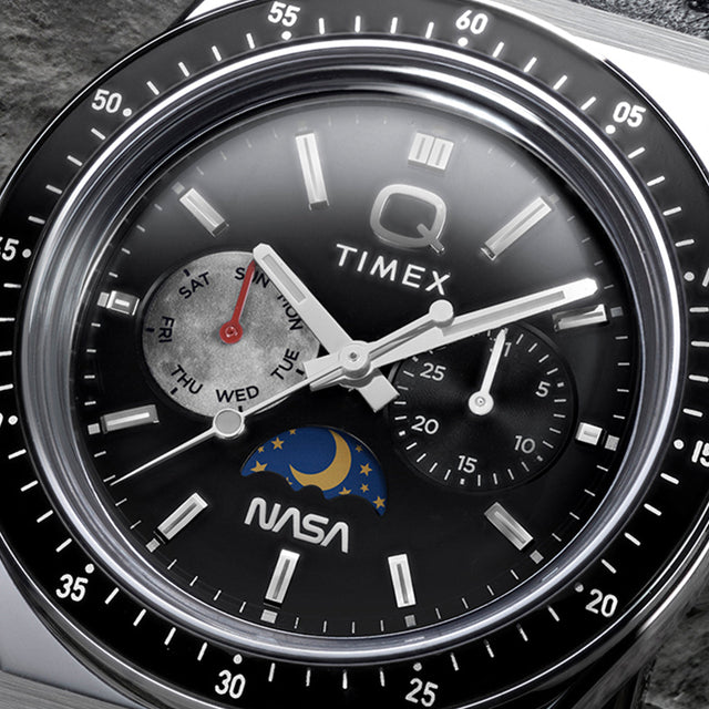 Close-up of Q Timex® x NASA black dial featuring day-of-week subdial with red hand, secondary subdial, moonphase detail, luminous markers, and NASA logo.