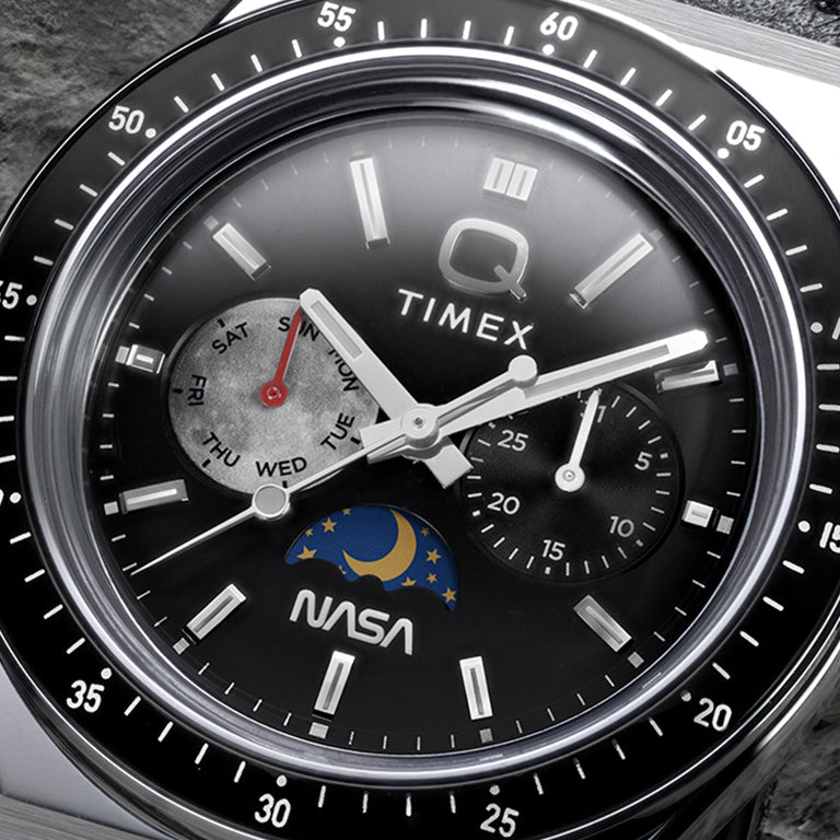 Close-up of Q Timex® x NASA black dial featuring day-of-week subdial with red hand, secondary subdial, moonphase detail, luminous markers, and NASA logo.