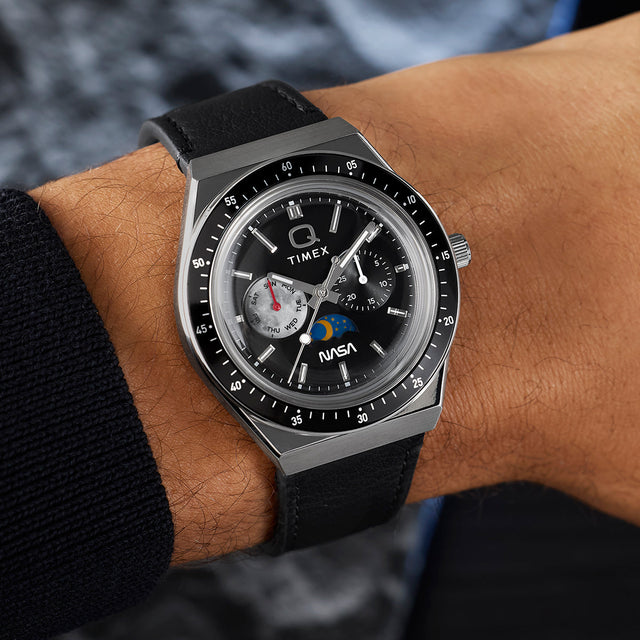 Q Timex® x NASA watch worn on wrist, showcasing brushed stainless steel case, black bezel, black leather strap, and multifunction dial with NASA logo.