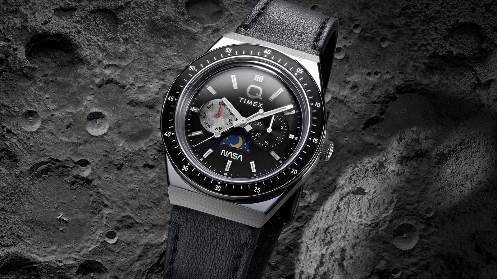 Angled view of Q Timex® x NASA watch with black leather strap set against textured moon-inspired surface.
