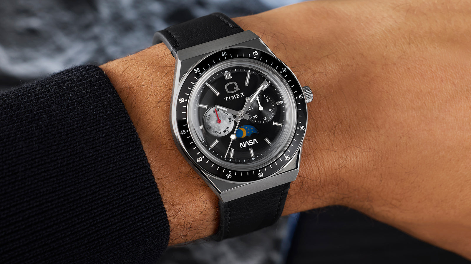 Close wrist shot of Q Timex® x NASA watch with black leather strap and black bezel against lunar-textured backdrop.