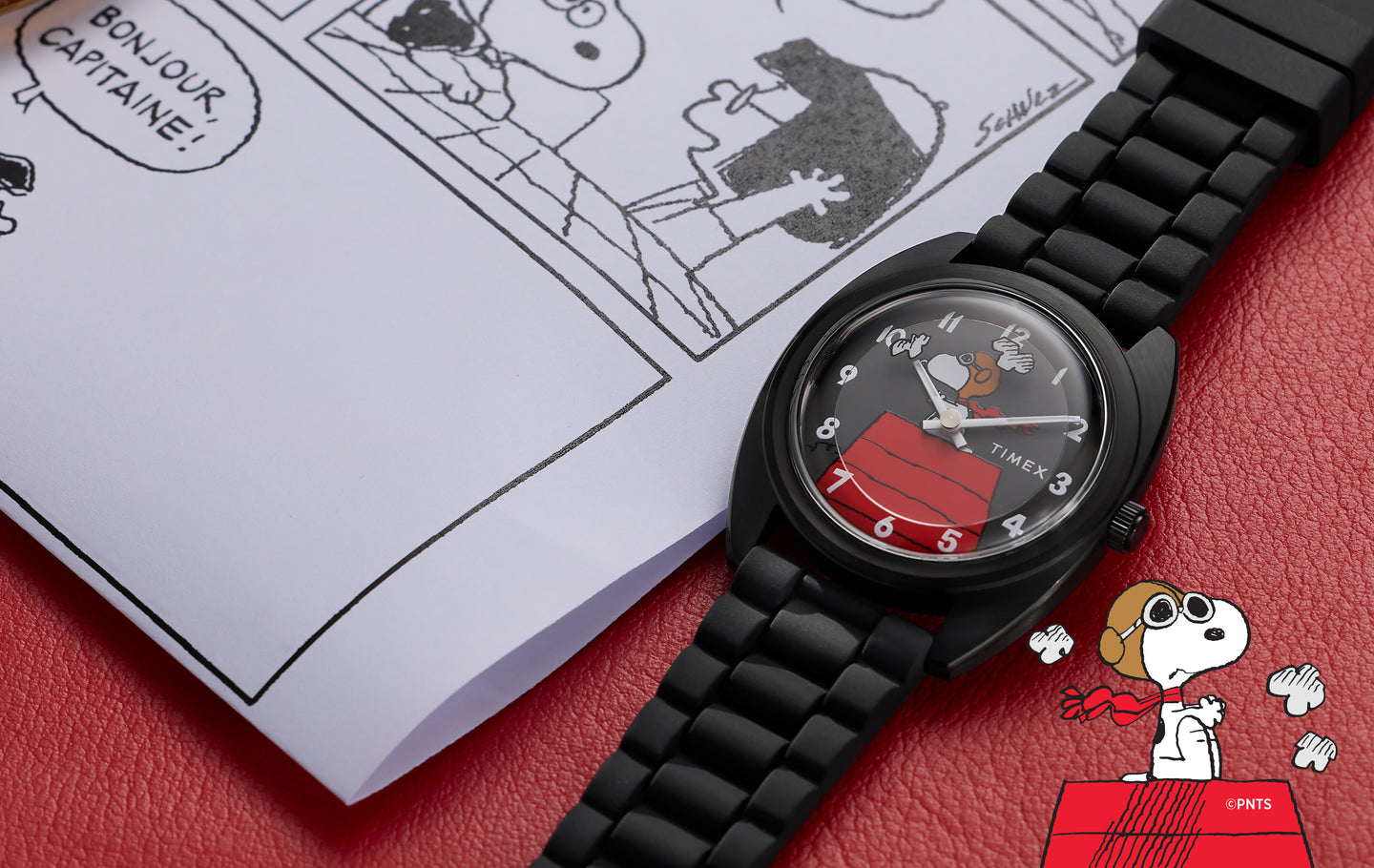 Peanuts® x Timex 1979 Reissue watch with a black case and bracelet, featuring Snoopy flying his red doghouse on the dial, placed on a red surface beside a Peanuts comic strip page.