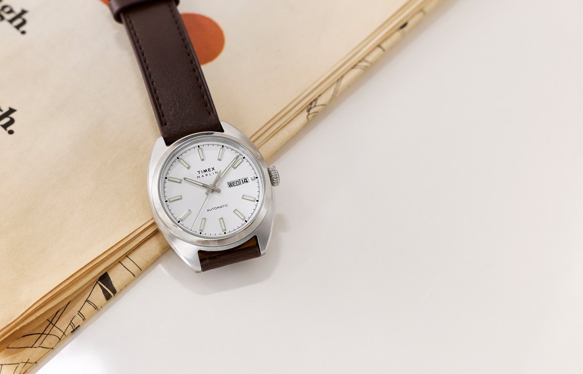 Timex Marlin® Draper Automatic watch with a brown leather strap resting on an open book, featuring a white dial and stainless steel case.