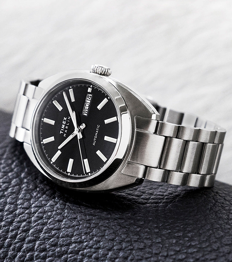 Timex Marlin Draper Automatic stainless steel watch with a black dial and day-date display, resting on a textured leather surface.