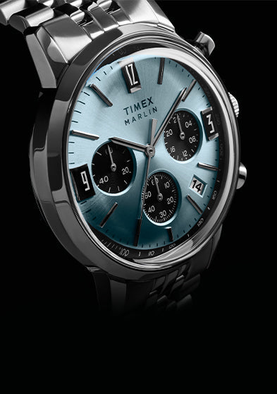 Timex Marlin Chronograph Tachymeter watch with a light blue dial, black subdials, tachymeter bezel, and stainless steel bracelet, shown at an angled close-up against a dark background.