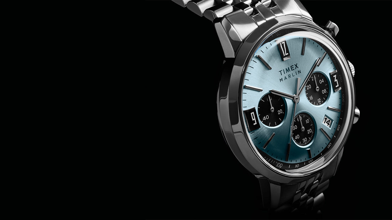 Timex Marlin Chronograph Tachymeter watch with a light blue dial, black subdials, tachymeter bezel, and stainless steel bracelet, shown at an angled close-up against a dark background.