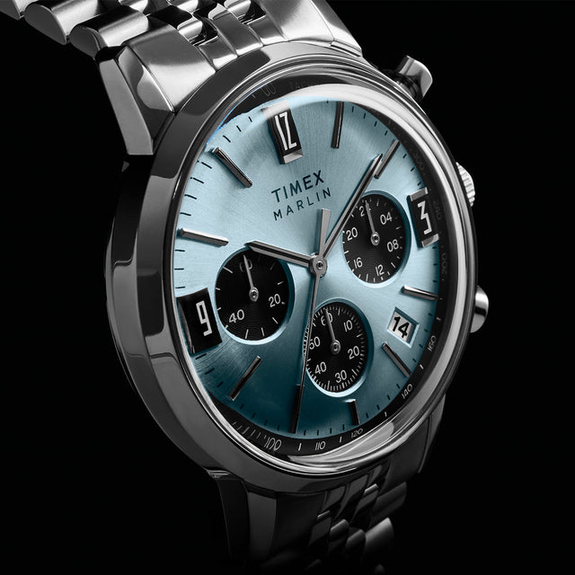 Timex Marlin Chronograph Tachymeter watch with a light blue dial, black subdials, tachymeter bezel, and stainless steel bracelet, shown at an angled close-up against a dark background.